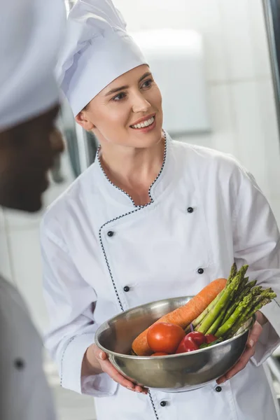 Chef sorridente che tiene la ciotola con le verdure e distoglie lo sguardo alla cucina del ristorante — Foto stock