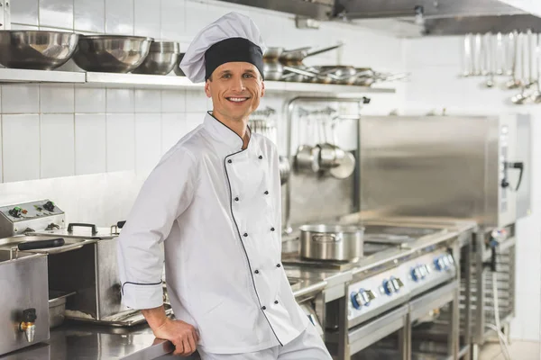 Smiling chef leaning on table and looking at camera at restaurant kitchen — Stock Photo