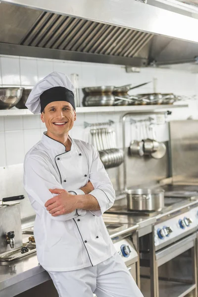 Chef sorridente in piedi con le braccia incrociate e guardando la fotocamera alla cucina del ristorante — Foto stock