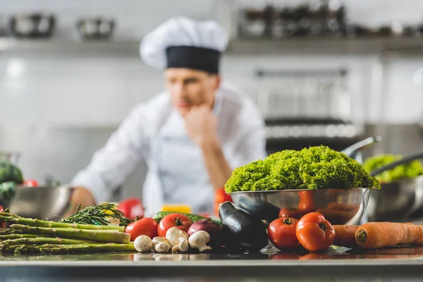Pensive chef at restaurant kitchen with vegetables on foreground — Stock Photo