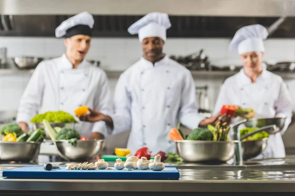 Chefs multiculturais na cozinha do restaurante com legumes em primeiro plano — Fotografia de Stock