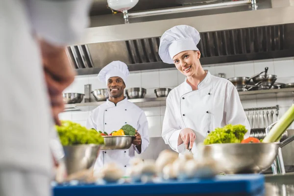 Cozinheiros multiculturais sorridentes preparando comida na cozinha do restaurante e olhando para a câmera — Fotografia de Stock