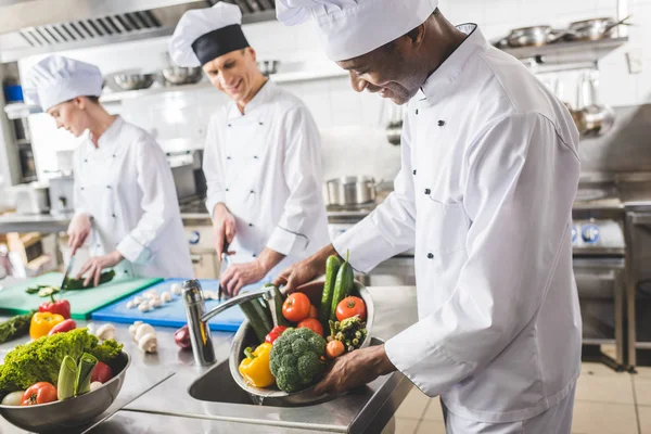 Sonriente afroamericano chef lavar verduras en la cocina del restaurante - foto de stock