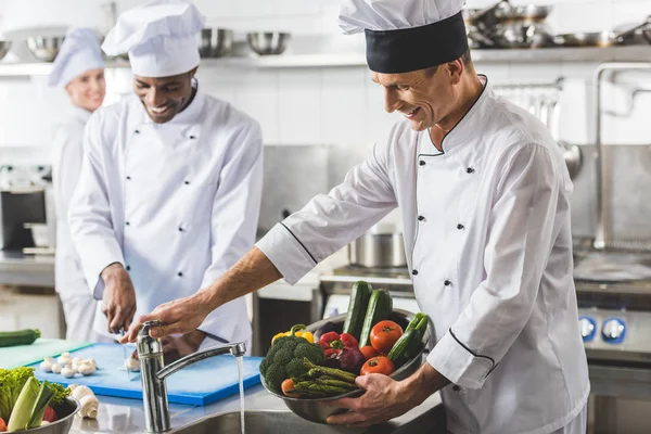 Chefs multiculturales cortando y lavando verduras en la cocina del restaurante - foto de stock