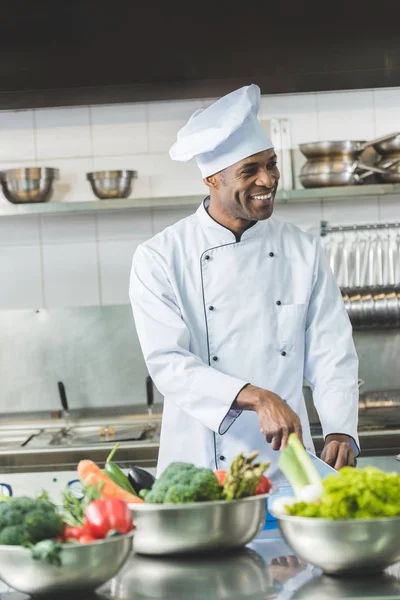 Sonriente guapo afroamericano chef cortar verduras en restaurante cocina - foto de stock