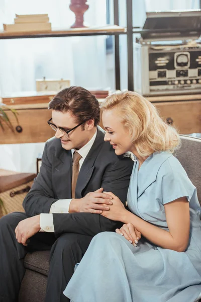Feliz pareja de estilo de los años cincuenta mirando hacia otro lado mientras está sentado en el sofá y cogido de la mano - foto de stock