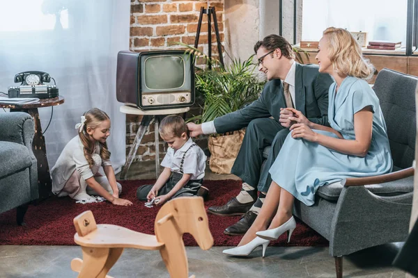 Padres felices sentados en el sofá y mirando a los niños pequeños jugando con fichas de dominó en casa, estilo de los años 50 - foto de stock