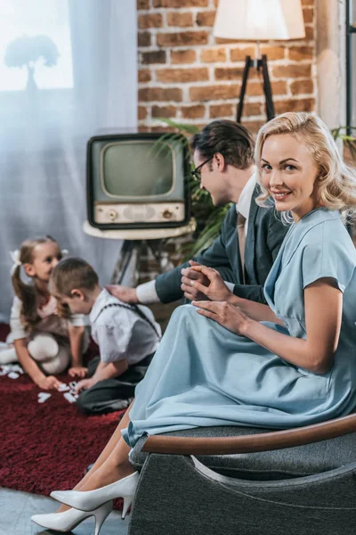 Mujer feliz estilo años 50 sonriendo a la cámara mientras se sienta en el sofá con el marido y los niños jugando con fichas de dominó - foto de stock