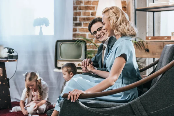 Glückliche altmodische Eltern, die auf dem Sofa sitzen und Händchen halten, während Kinder mit Domino-Fliesen spielen, Stil der 1950er Jahre — Stockfoto