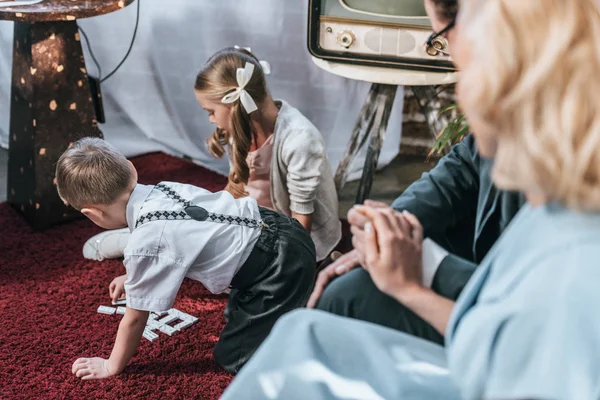 Cropped shot of parents sitting on sofa and holding hands while children playing with domino tiles, 1950s style — Stock Photo