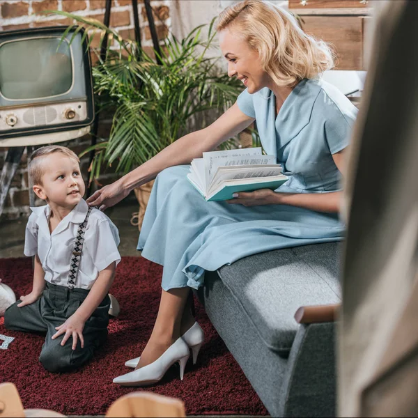 Счастливая блондинка читает книгу и смотрит на симпатичного маленького сына, играющего дома на ковре, семья в стиле 1950-х годов — стоковое фото