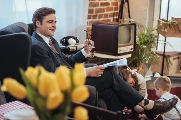 Lächelnder Mann im Anzug, der im Sessel Zeitung liest, während Kinder im Hintergrund mit Domino-Fliesen spielen, Familie im Stil der 1950er Jahre — Stockfoto