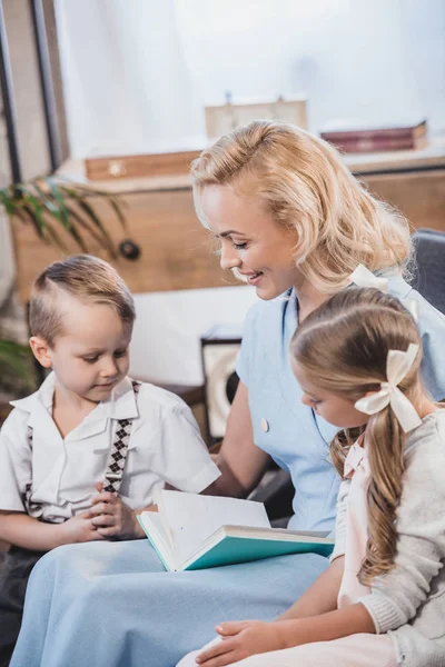 Happy mother with adorable little kids reading book together at home, 1950s style family — Stock Photo