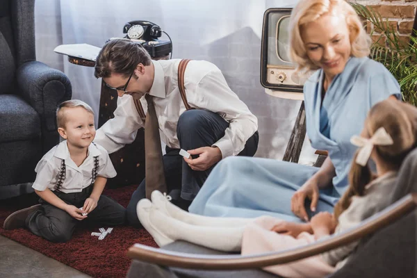 Feliz madre e hija sentadas en el sofá, mientras sonríen padre e hijo jugando con fichas de dominó, estilo de los años 50 - foto de stock