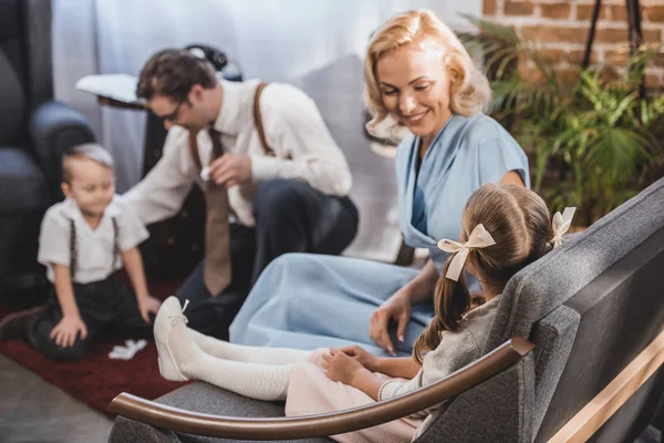 Happy mother looking at cute little daughter sitting on sofa while father with son playing dominoes at home, 1950s style — Stock Photo