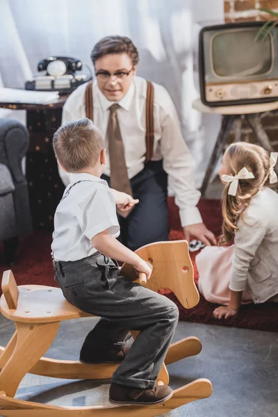 Mignon petit garçon assis sur cheval à bascule et regardant père jouer dominos avec fille, famille de style des années 50 — Photo de stock