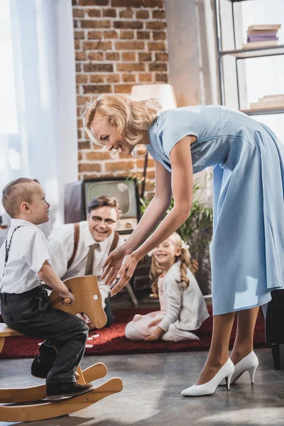 Happy mother, father and sister looking at cute little boy sitting on rocking horse at home, 50s style — Stock Photo