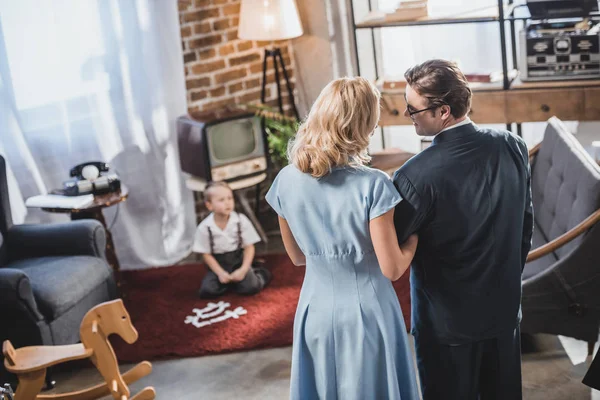 Rückseite: Eltern stehen zusammen und lächeln sich an, während ihr süßer kleiner Sohn mit Dominosteinen spielt, Stil der 1950er Jahre — Stockfoto