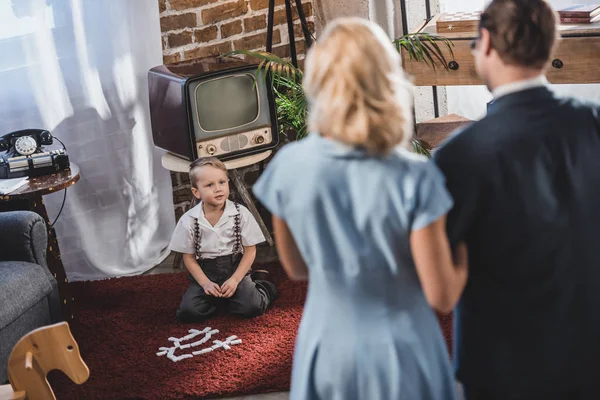 Вид сзади родителей, стоящих вместе и смотрящих на симпатичного маленького сына, играющего с плиткой в стиле 1950-х годов — стоковое фото