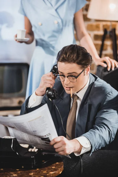 Uomo con giornale di lettura sigaretta e parlando per telefono vintage mentre la donna tiene una tazza di caffè dietro — Foto stock