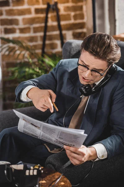 Emotional man in suit and eyeglasses pointing at newspaper and talking by vintage telephone — Stock Photo