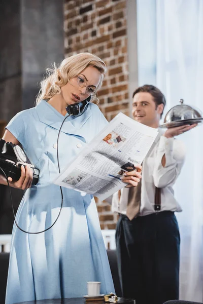 Serious woman reading newspaper and talking by vintage telephone while husband holding meal behind, 50s style — Stock Photo