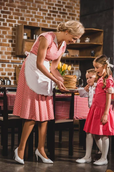 Feliz hermano y hermana mirando deliciosos panqueques mientras la madre sostiene la placa, familia de estilo de los años 50 - foto de stock