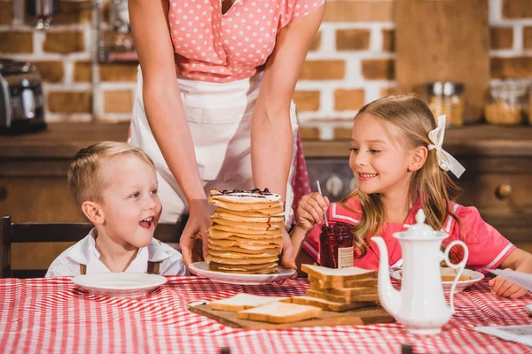 Plan recadré de mère mettant des crêpes sur la table et regardant des enfants souriants mignons prendre le petit déjeuner, famille de style des années 50 — Photo de stock