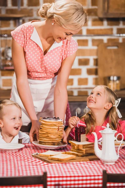 Mère heureuse mettant des crêpes sur la table et regardant des enfants souriants mignons prendre le petit déjeuner, famille de style des années 50 — Photo de stock