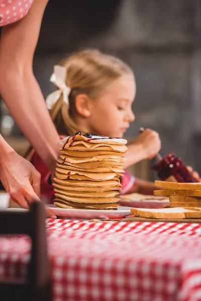 Cropped shot of woman holding plate with pancakes and child eating jam behind — Stock Photo