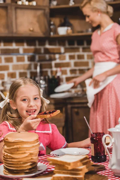 Adorable niña comiendo tostadas con mermelada y sonriendo a la cámara mientras la madre lava los platos detrás, estilo de los años 50 - foto de stock