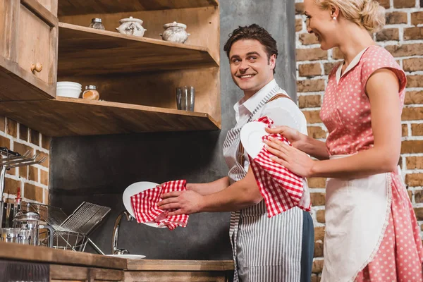 Coppia in grembiuli che si sorridono mentre lavano i piatti insieme, stile anni '50 — Foto stock