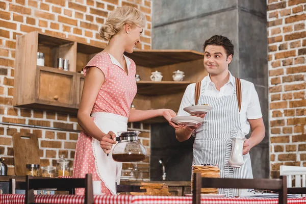 Mann und Frau in Schürzen lächeln einander beim Servieren des Frühstückstisches im Stil der 50er Jahre zu — Stockfoto