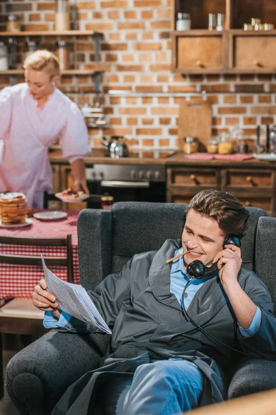 Smiling man in robe reading newspaper, smoking cigarette and talking by vintage telephone while wife preparing breakfast behind — Stock Photo