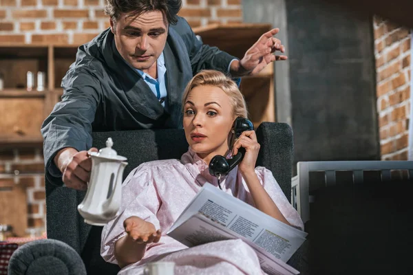 Жінка в халаті читає газету і розмовляє по телефону, а чоловік тримає каструлю, стиль 1950-х років — стокове фото