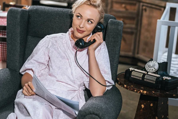 Sonriente mujer rubia en bata sosteniendo periódico y hablando por teléfono vintage en casa - foto de stock