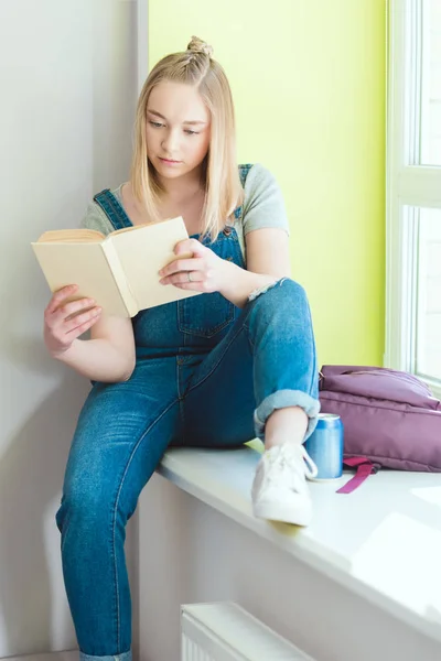 Teenage schoolgirl reading book and sitting on window sill with soda and backpack — Stock Photo