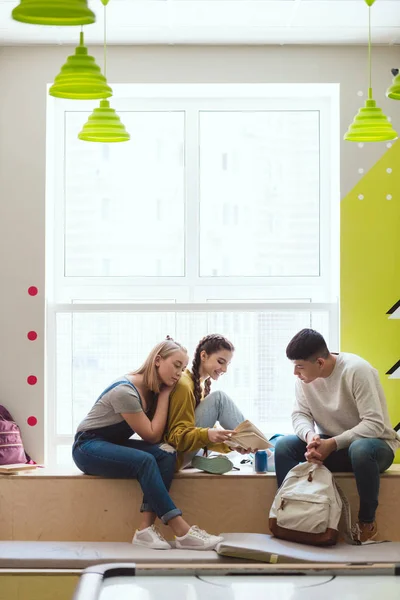 Multicultural group of teenage schoolchildren sitting on window sill and reading book — Stock Photo