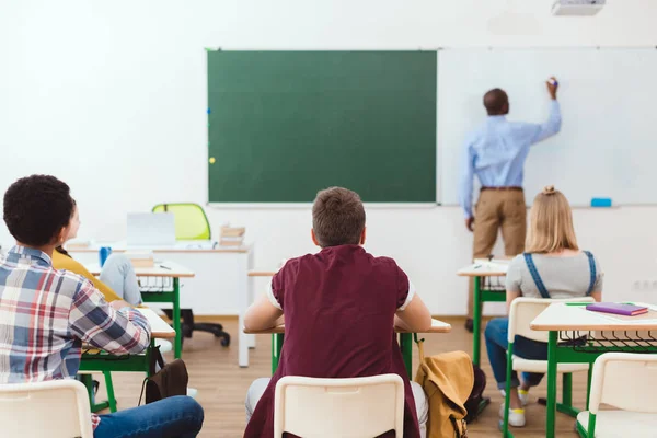 Vista trasera de los escolares y la escritura del profesor en pizarra blanca - foto de stock