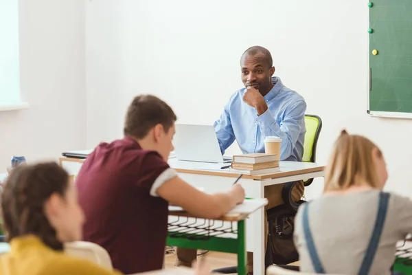 Escrevendo escolares e professores sentados à mesa com laptop e xícara de café — Fotografia de Stock