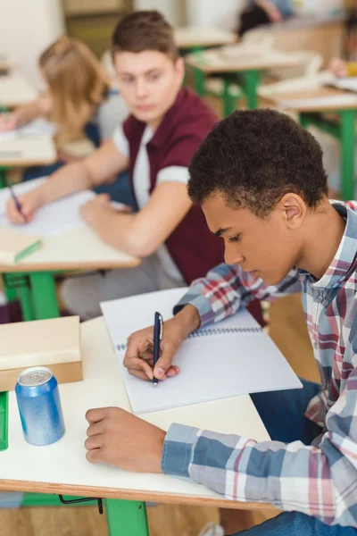 Elevata vista di afro-americano adolescente scolaro scrivere in un libro di testo e compagni di classe dietro — Foto stock