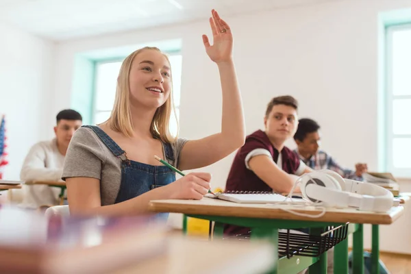 Colegial adolescente estudante com braço para cima e colegas sentados atrás em sala de aula — Fotografia de Stock