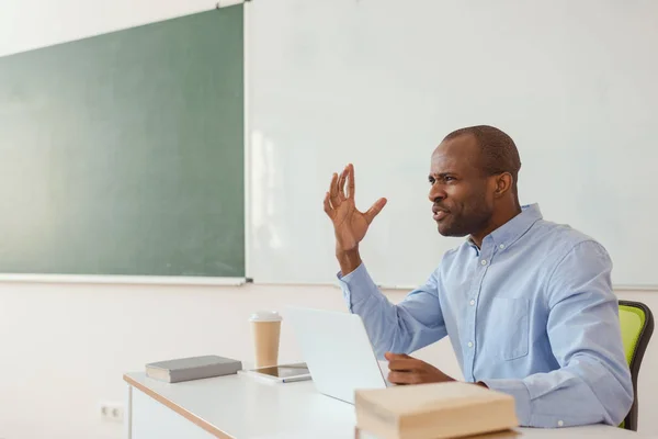 Frustrado profesor afroamericano haciendo gestos y sentado en el escritorio con el ordenador portátil - foto de stock