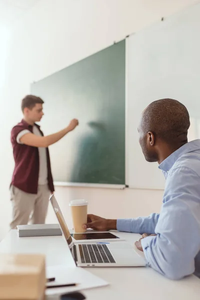 High school student writing at chalk board and african american teacher sitting at desk with laptop and coffee cup — Stock Photo