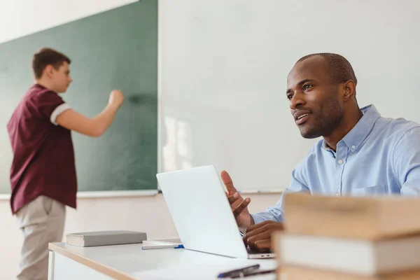 Lycée adolescent écriture sur tableau noir et professeur afro-américain assis au bureau avec ordinateur portable et expliquer — Photo de stock