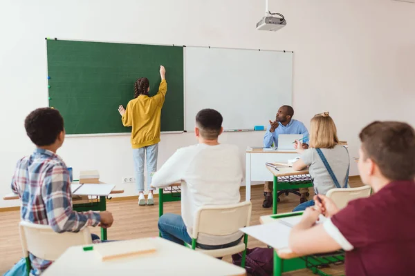 Rear view of schoolgirl writing on chalk board and classmates with teacher sitting in classroom — Stock Photo