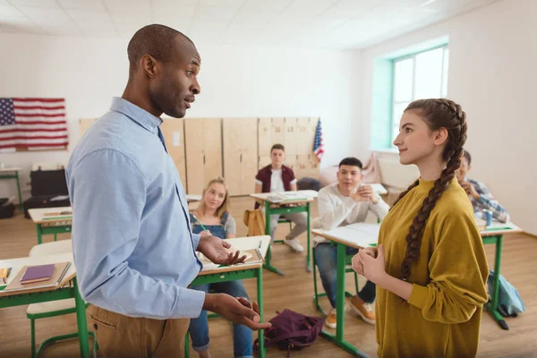 Vista lateral del profesor afroamericano haciendo gestos y hablando con la colegiala con compañeros sentados detrás - foto de stock