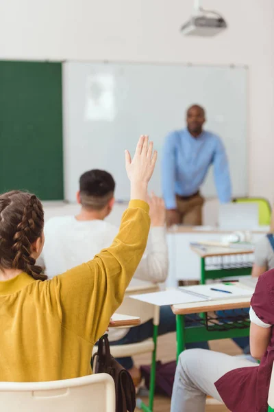 Rückansicht einer Schülerin mit erhobenem Arm mit Klassenkameraden und Lehrer im Klassenzimmer — Stockfoto