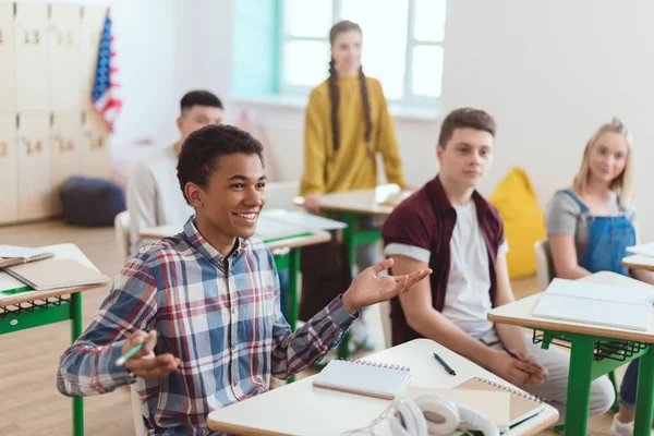 Un garçon afro-américain souriant aux bras larges assis à table en classe avec des écoliers — Photo de stock