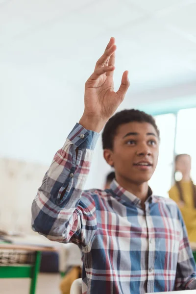 Estudiante adolescente afroamericano con el brazo en alto y compañeros de clase detrás en el aula - foto de stock